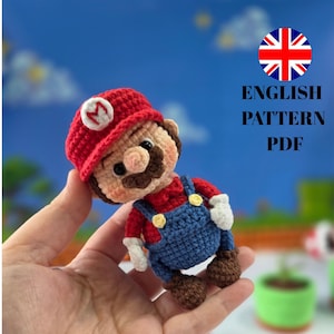 Puede incluir: Muñeco de Mario tejido a ganchillo, con gorra roja, bigote marrón y peto azul. El muñeco se sostiene en una mano, con el texto 'ENGLISH PATTERN PDF' visible en el fondo.