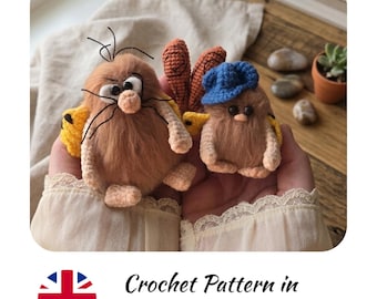 May include: Two fuzzy, light brown crochet creatures with small black eyes and light peach-colored feet. The larger creature has thin black whiskers and yellow wings, while the smaller one wears a blue crochet hat and has yellow wings.