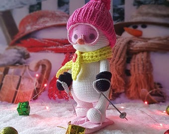 Puede incluir: Un muñeco de nieve de ganchillo con gorro rosa, gafas y bufanda amarilla, sosteniendo bastones de esquí y de pie sobre esquís rosas. El muñeco de nieve tiene mitones negros y una pequeña caja de regalo dorada a sus pies. El fondo presenta otros muñecos de nieve y un paisaje nevado.