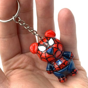 Puede incluir: Un llavero de oso de peluche rojo y azul con un diseño de Spiderman. El oso lleva un traje rojo y azul con un patrón de telaraña. El llavero tiene un anillo de metal plateado.