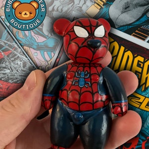 May include: A red and blue Spider-Man themed teddy bear with a black web design on the chest. The bear is holding its hands in front of its body. The bear is sitting on a hand.
