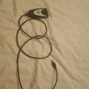 May include: A black and silver digital converter box with a black cable and a USB connector. The converter box is labeled "MINI" and has the brand name "AVerMedia" printed on it. The cable is coiled on a white quilted surface.