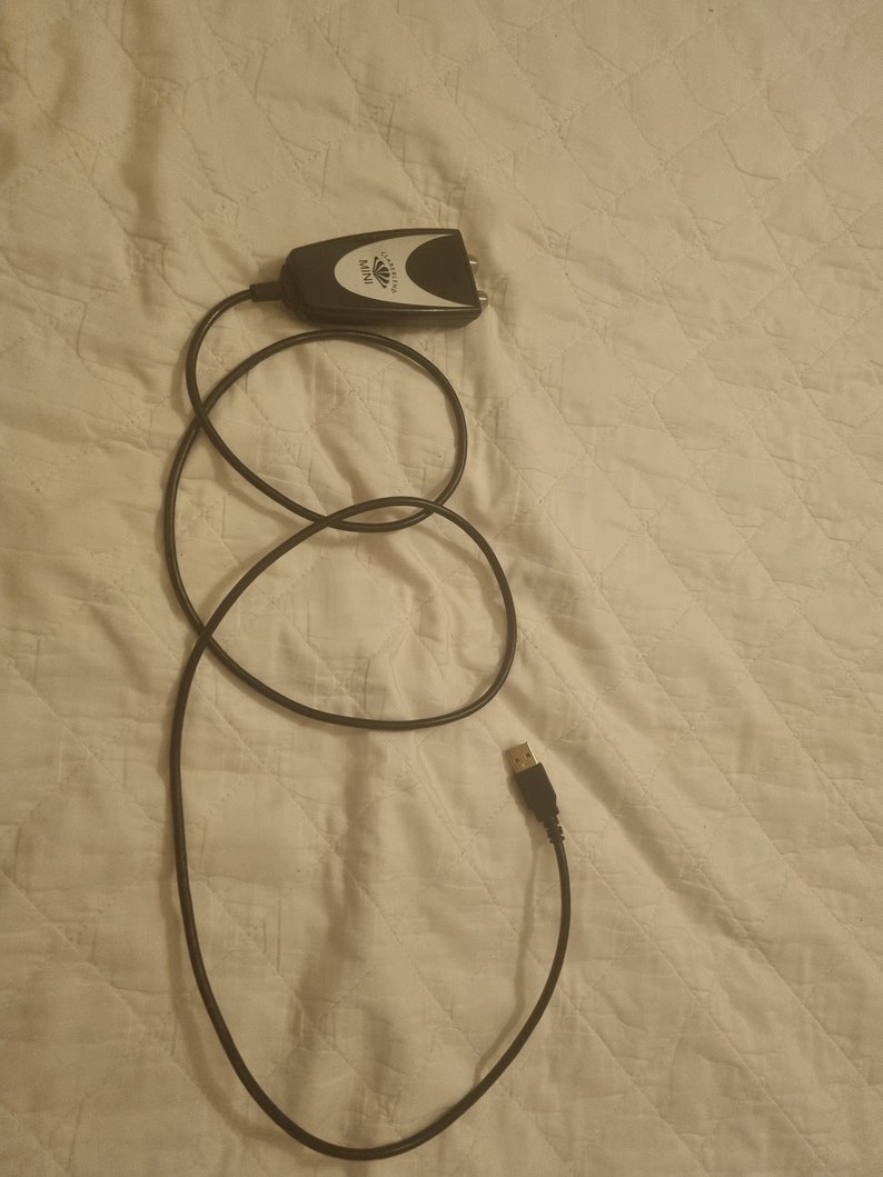 K&ouml;nnte beinhalten: Ein schwarzer und silberner digitaler Konverter mit einem schwarzen Kabel und einem USB-Anschluss. Der Konverter ist mit "MINI" beschriftet und tr&auml;gt den Markennamen "AVerMedia". Das Kabel ist auf einer wei&szlig;en, gesteppten Oberfl&auml;che aufgerollt.