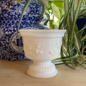 May include: A white, pedestal planter with embossed floral detailing. The planter has a wide, slightly flared rim and a decorative, ribbed base. The background includes greenery and a blue and white patterned ceramic piece.