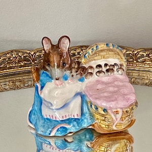 Peut inclure: Une figurine en porcelaine d'une souris brune vêtue de bleu et de rose, tenant un panier avec une couverture rose et plusieurs petites souris à l'intérieur.