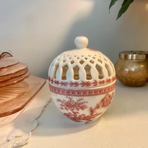 Peut inclure: Un pot en céramique blanche de forme sphérique avec un couvercle amovible. Le couvercle présente un motif ajouré en treillis. Le corps est orné d'un motif rouge représentant des arbres et des personnages, avec une bordure florale au milieu.