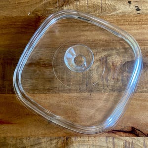 May include: A square, clear glass lid with rounded corners and a central, circular knob handle. The lid features a smooth, transparent surface with a slightly raised rim designed to fit securely onto a corresponding square container or dish.