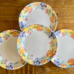 May include: Three round ceramic plates featuring a white centre and a wide yellow border decorated with a colourful pattern of painted peaches, cherries, and clusters of purple grapes arranged around the rim of each individual dinner plate.