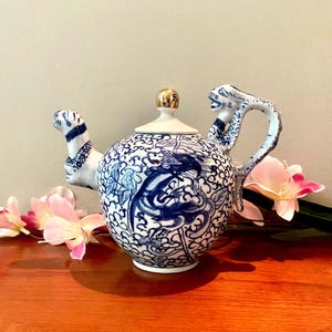 May include: A ceramic teapot with a blue and white dragon design. The teapot has a gold-coloured lid and a dragon-shaped handle. The teapot is decorated with blue floral patterns.
