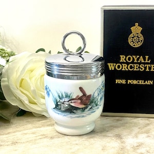 Peut inclure: Un coquetier en porcelaine blanche avec un couvercle et une poignée argentés. Le coquetier présente une scène peinte d'un oiseau brun avec un long bec, entouré de feuillage bleu et vert. La boîte indique "Royal Worcester Fine Porcelain".