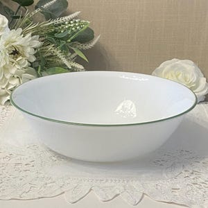 Peut inclure: Un bol blanc avec un bord vert. Le bol est rond et repose sur une nappe en dentelle blanche. Des fleurs blanches et de la verdure sont en arrière-plan.