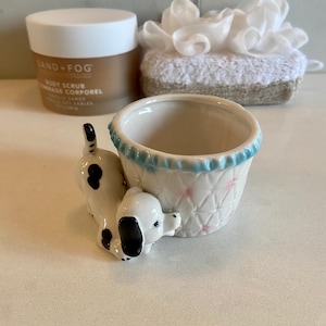 May include: A small ceramic planter featuring a white dog with black spots attached to the side. The planter has a white basket-weave texture with small pink accents and a light blue rim. Behind it sits a Sand + Fog body scrub jar and a white loofah.