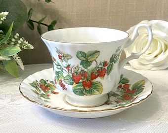 May include: A white teacup and saucer set featuring a strawberry pattern. The teacup and saucer are adorned with red strawberries, green leaves, and gold edging. The teacup has a curved handle and rests on a matching saucer.