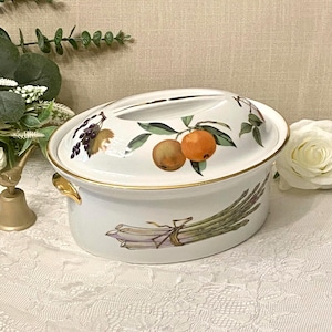 May include: An oval white ceramic casserole dish with a lid, featuring a gold-trimmed rim and gold-toned side handles. The exterior displays a painted illustration of a bundle of green asparagus, while the lid shows painted fruit and leaves.