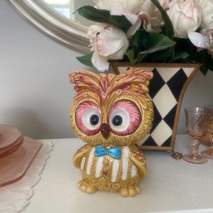 May include: A ceramic owl figurine with a yellow and white striped shirt, a blue bow tie, and large pink eyes. The owl is standing on a white surface.