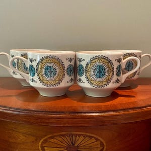 May include: Four white ceramic coffee mugs with a decorative design. Each mug features a central teal and gold circular pattern, surrounded by a border of gold and teal accents. The mugs have a classic handle design.