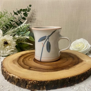 Peut inclure: Une tasse en céramique crème avec un bord rose et une anse, ornée d'un motif floral bleu et rose. La tasse est posée sur une tranche de bois ronde.