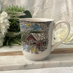 May include: Cream ceramic mug with a detailed illustration of a covered bridge scene. The mug has a curved handle and a brown rim. The artwork includes a red bridge, trees, and water, with a blue and yellow sky.