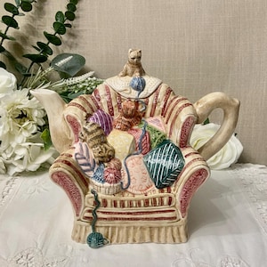 May include: A decorative ceramic teapot in the shape of an armchair, featuring beige and red striped patterns. The chair is filled with colourful cushions and small cat figurines. A small cat sits on the lid, and a ball of yarn is at the base.