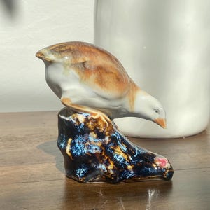 May include: A porcelain figurine of a bird with brown and white plumage perched on a dark, textured base with blue and black mottled glaze. The bird is depicted in a forward-leaning posture with a small orange beak and a smooth, glossy finish.