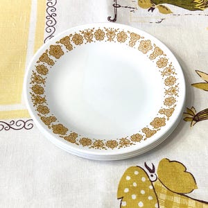 May include: A stack of white ceramic plates with a gold floral pattern around the rim. The plates are stacked on a patterned tablecloth with yellow and white floral designs.