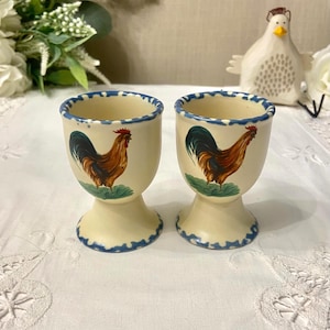 May include: Two cream-coloured ceramic egg cups with a blue scalloped rim and base. Each cup features a detailed illustration of a rooster in shades of brown, red, and green. A decorative chicken figurine is in the background.