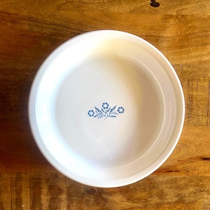 May include: A white ceramic pie dish with a blue floral pattern in the centre. The dish has a slightly raised rim. The floral design features small blue flowers and leaves. It is set against a wooden surface.