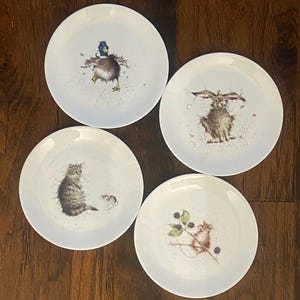 May include: Four white ceramic plates, each with a different animal illustration. A duck, rabbit, cat and mouse, and a mouse with berries are depicted. The plates are displayed on a wooden surface.