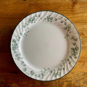May include: White ceramic dinner plate with a green ivy leaf pattern around the edge. The plate has a green border and a ridged edge. The plate is on a wooden surface.