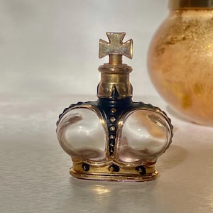 May include: A crown-shaped perfume bottle with a gold base and accents, a black band, and a cross finial. The bottle has a pearlescent, rounded body. A gold-coloured jar is visible in the background.