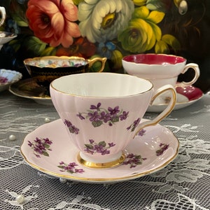 Peut inclure: Une tasse à thé et une soucoupe roses avec un motif floral délicat de violettes violettes. La tasse à thé a un design cannelé et un bord doré. La soucoupe a un motif floral assorti.