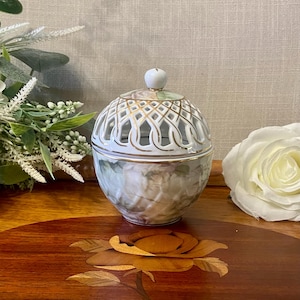 May include: A round porcelain jar with a lid featuring a white and gold lattice pattern. The body of the jar is decorated with a soft, muted floral print in shades of pink, green, and cream. The lid is topped with a small, smooth white knob.