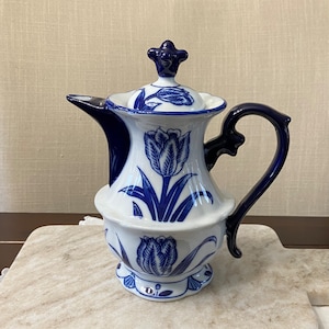 May include: A white and blue ceramic pitcher featuring a tulip design. The pitcher has a dark blue handle and spout, and a matching lid with a decorative finial. The tulip design is repeated around the body of the pitcher.