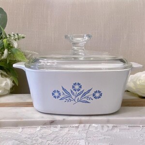 May include: A white rectangular glass casserole dish with a clear glass lid and a blue floral pattern on the front. The dish features integrated side handles and a central knob on the lid, showcasing a symmetrical design of blue flowers and leaves.