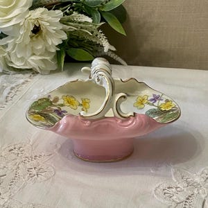 May include: A vintage pink porcelain basket with a decorative handle and floral design. The basket has a scalloped edge with yellow and purple flowers. The handle and trim are gold-coloured.
