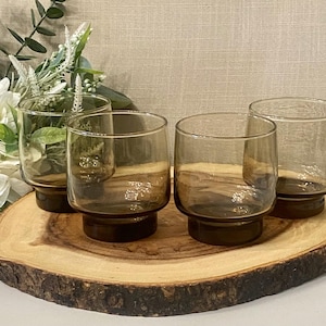 May include: Four amber-tinted, short drinking glasses with a rounded base. The glasses are arranged on a round, wooden serving board. The background includes white flowers and greenery.