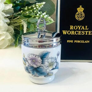 May include: A white porcelain egg coddler featuring a floral pattern with blue and purple flowers and green leaves. The container has a polished silver-tone metal lid with a circular ring handle at the top, positioned next to a black box with gold text.