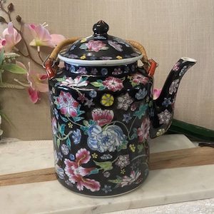 May include: A black ceramic teapot featuring a vibrant floral pattern with pink, blue, white, and yellow flowers. The teapot has a matching lid, a curved spout, and a curved handle made of light brown woven material attached to orange metal loops.