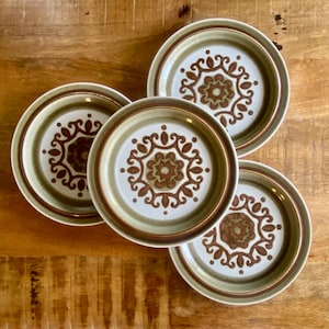 May include: A set of four round ceramic plates featuring a white centre with a brown and olive green floral medallion pattern. Each plate has a wide, olive green rim and a smooth, glazed finish, arranged in a cluster to display the circular design.