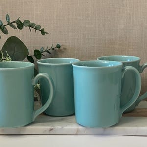 May include: Four pale blue ceramic mugs with curved handles sit on a white marble tray. The mugs are cylindrical with a slightly flared rim. Greenery and a decorative bird are in the background.