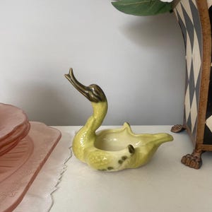 May include: A ceramic swan-shaped trinket dish with a green glaze and brown accents. The dish has a small, round opening in the centre.