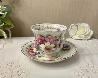 May include: A floral teacup and saucer set. The teacup and saucer are white with a gold trim. The teacup has a handle and is decorated with pink and white flowers. The word "October" is printed on the teacup.