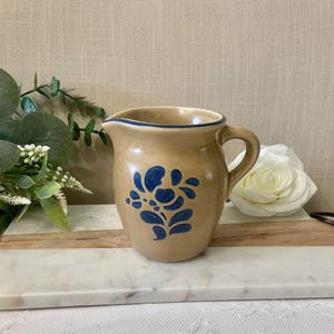 May include: A tan ceramic pitcher featuring a dark blue floral pattern on the front, a curved handle, and a dark blue rim. The pitcher has a smooth, glossy finish and a rounded body shape with a small pouring spout at the top edge.