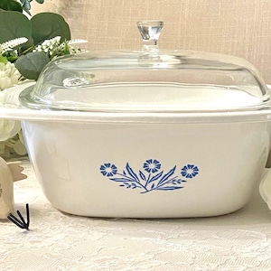 May include: A white oval casserole dish with a clear glass lid, featuring a blue floral pattern on the front. The dish has integrated side handles and a smooth finish. A small decorative chicken figurine and white flowers are placed nearby.