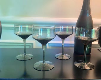 May include: A set of four smoke-gray glass coupe glasses with wide, shallow bowls and slender stems. Each glass features a translucent, dark-tinted finish and a circular base. The glasses are arranged in a row next to a dark wine bottle.