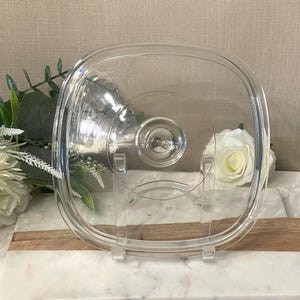 May include: A clear, square-shaped glass lid with rounded corners and a central circular handle. The lid features a smooth, transparent surface and a raised rim, designed to fit securely onto a corresponding square container or cookware piece.