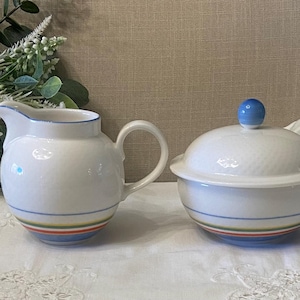 May include: A white ceramic creamer and lidded sugar bowl set. The creamer has a handle and spout, while the sugar bowl has a blue knob on the lid. Both pieces have blue trim and colourful stripes around the base. Decorative floral arrangement in the background.