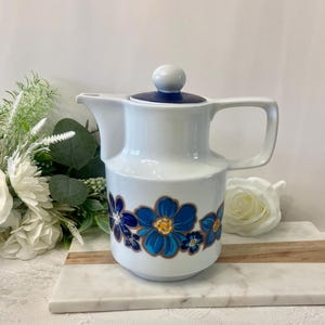 Peut inclure: Pichet en céramique blanche avec un motif floral bleu et or. Le pichet a une poignée et un couvercle avec un dessus bleu et un bouton blanc. Le motif floral présente des fleurs bleues avec des centres dorés et des contours bruns. Un article décoratif pour la cuisine.
