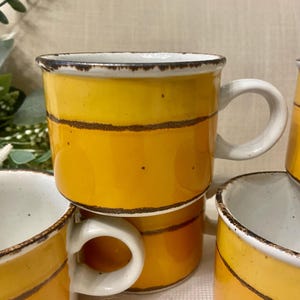 May include: A set of ceramic mugs featuring a bright yellow and orange horizontal striped pattern with dark brown, distressed-look rim and stripe accents. Each mug has a white interior and a sturdy, curved white handle, showcasing a speckled finish.