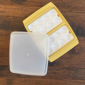 May include: A yellow plastic egg container with a clear lid. The container is divided into two sections, each holding 6 egg-shaped indentations. The lid is off to the side, revealing the white interior. The container is on a wooden surface.